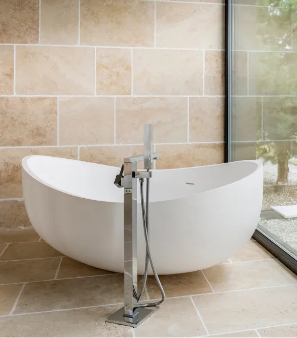 Pacific Designer Bathrooms offers a modern freestanding white bathtub with a sleek silver faucet, set against a tiled wall and under a large window overlooking greenery.