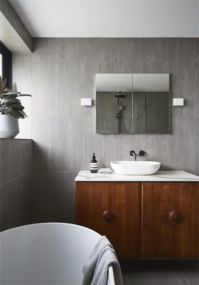 Modern bathroom by Pacific Designer Bathrooms with wooden vanity cabinet, white basin, and wall-mounted mirror. Dark gray tiles cover the walls and floor. A white bathtub and a potted plant are visible.