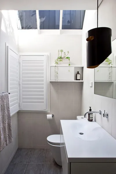 Modern bathroom by Pacific Designer Bathrooms featuring a white basin and toilet, gray floor tiles, a large hanging black pendant light, a shuttered window and skylight, with green plants on a shelf